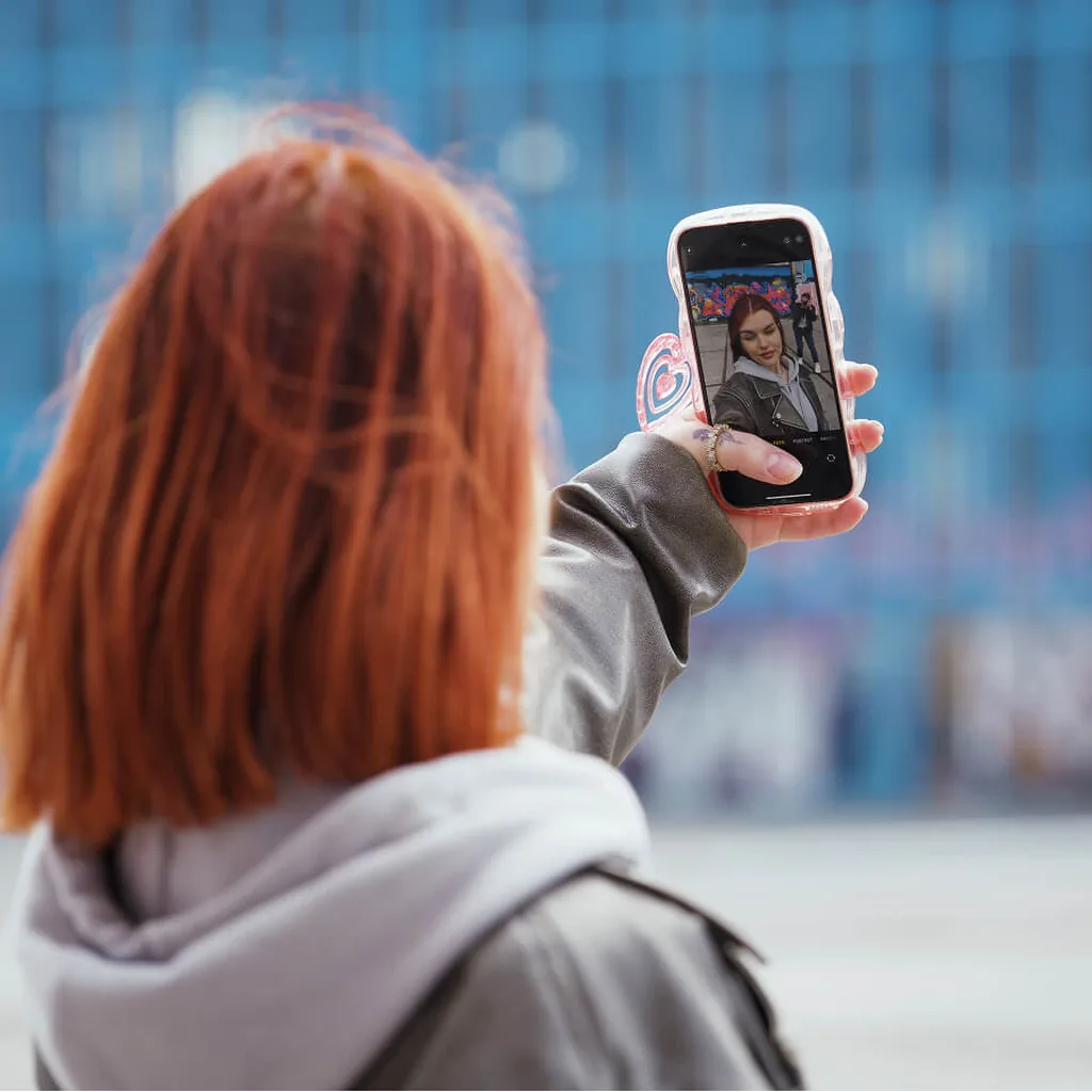 A woman with red hair takes a selfie on her phone in front of a blue building.